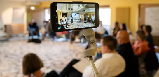 Ein Smartphone auf einem Stativ zeichnet ein Meeting oder einen Workshop auf. Die Teilnehmer sitzen im Kreis in einem hell erleuchteten Raum. Der Fokus liegt auf dem Display des Smartphones, das die Gruppe und den Moderator im Vordergrund zeigt.