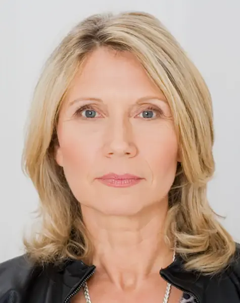 A middle-aged woman with shoulder-length blonde hair and blue eyes looks directly at the camera. She is wearing a black jacket and a light top, with a neutral facial expression against a plain background.