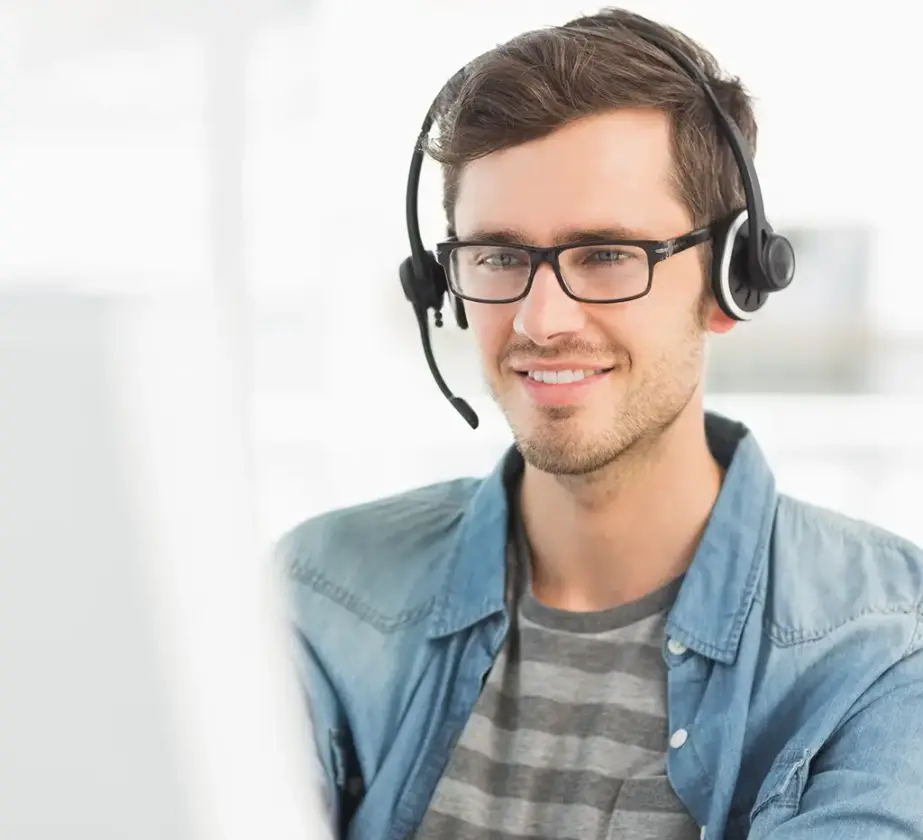 A young man wearing glasses and a headset smiles while looking at a computer screen. He is casually dressed in a striped shirt and denim jacket, working in a bright, modern office setting.