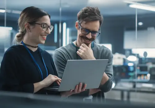 Zwei Kollegen mit Brillen stehen lächelnd in einem modernen Büro und schauen gemeinsam auf einen Laptop-Bildschirm, was eine Zusammenarbeit oder Diskussion an einem Projekt nahelegt.