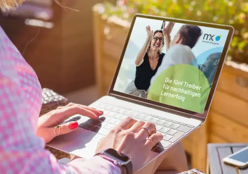 Eine Person, die im Freien einen Laptop benutzt, sieht einen Bildschirm, auf dem eine lächelnde Frau einem Mann ein High Five gibt. Der deutsche Text lautet: „Die fünf Treiber für nachhaltigen Lernerfolg“. Im Hintergrund sind bunte Blumen und Sonnenlicht zu sehen.