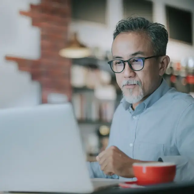 A middle-aged man with glasses and gray hair sits at a table using a laptop. He is wearing a light blue shirt and appears focused. A red coffee cup is on the table in front of him. The background shows a cozy, blurred café.