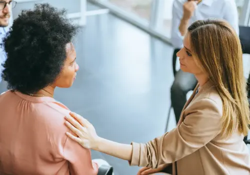 Eine Frau in einem beigen Blazer legt ihre Hand sanft auf die Schulter einer anderen Frau in einem rosa Oberteil und bietet Trost während eines Gruppentreffens in einem hellen, modernen Raum, in dem andere in der Nähe sitzen.
