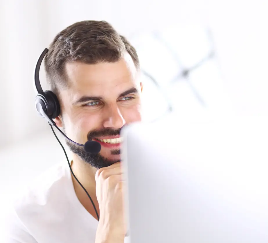 Ein lächelnder IT-Experte mit Bart und Headset sitzt vor einem Computer, wirkt engagiert und aufmerksam, was auf eine Kommunikation per Anruf oder Videochat in einer hellen, modernen Umgebung hindeutet.