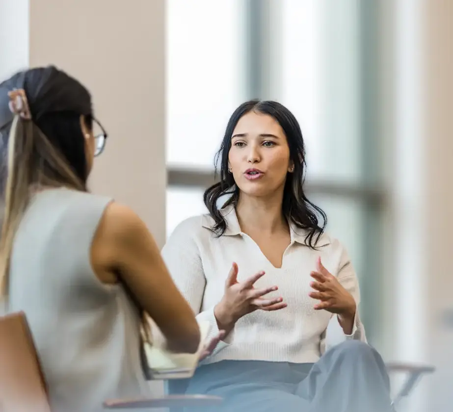 Zwei Frauen sitzen sich im Gespräch gegenüber und nehmen am Kommunikationscoaching teil. Die eine gestikuliert mit den Händen, die andere hört aufmerksam zu. Beide sitzen in einem hellen Raum mit großen Fenstern.