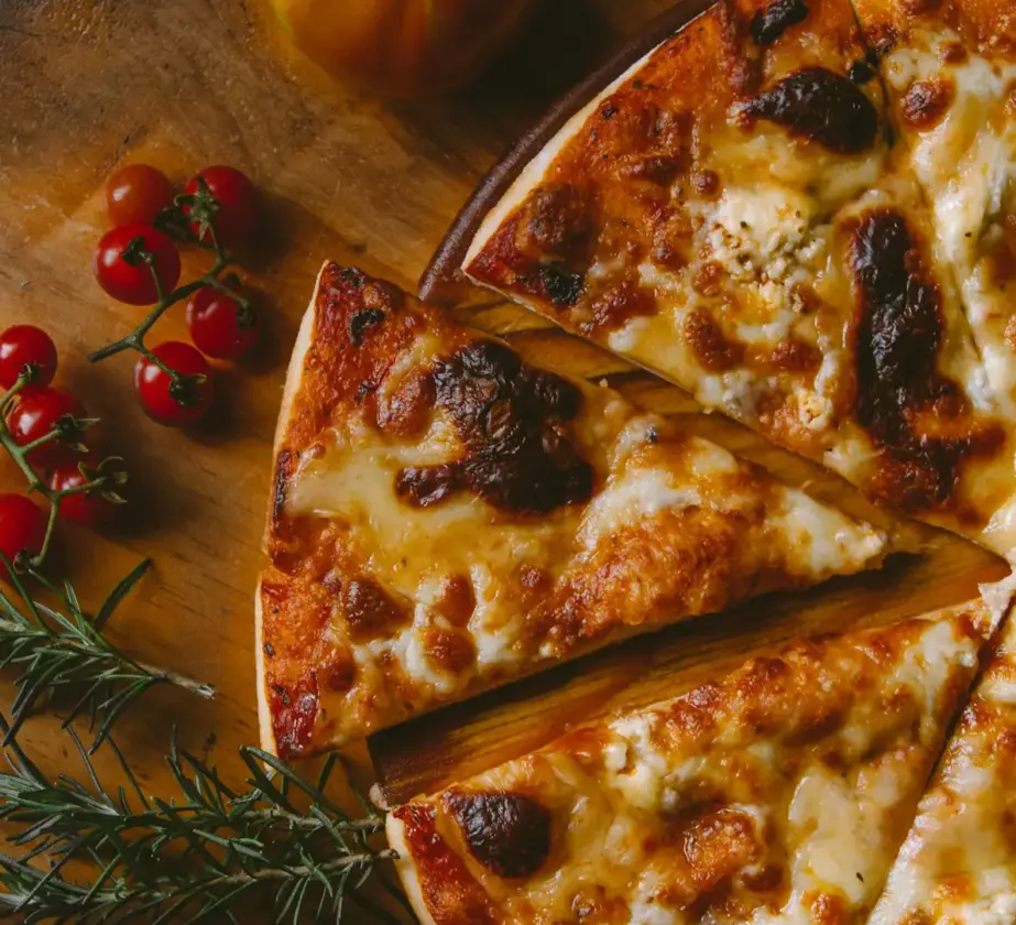 A cheese pizza sliced into pieces sits on a wooden surface, with fresh cherry tomatoes and sprigs of rosemary nearby.