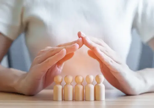 Eine Person in einem weißen Hemd formt mit ihren Händen über vier hölzernen menschlichen Figuren auf einem Tisch eine schützende Dachform und symbolisiert damit den Schutz oder die Fürsorge für Menschen oder eine Familie.