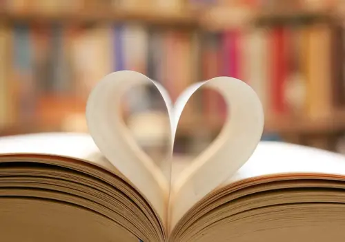 Ein offenes Buch mit zwei nach innen gebogenen Seiten, die eine Herzform bilden, vor einem unscharfen Hintergrund aus bunten Bücherregalen – perfekt für alle, die ihre Lernmotivation stärken möchten.