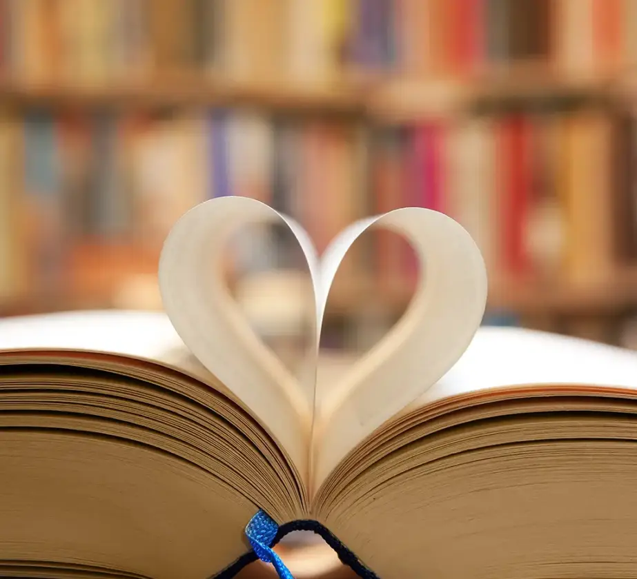 Ein offenes Buch mit zwei nach innen gebogenen Seiten, die eine Herzform bilden, vor einem unscharfen Hintergrund aus bunten Bücherregalen – perfekt für alle, die ihre Lernmotivation stärken möchten.