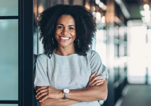 Eine Frau mit lockigem Haar, einem gestreiften Kleid und einer Uhr steht mit verschränkten Armen und einem selbstbewussten Lächeln in einem modernen, gut beleuchteten Büroflur - bereit, jede Herausforderung, vielleicht sogar einen Persönlichkeitstest, anzunehmen.