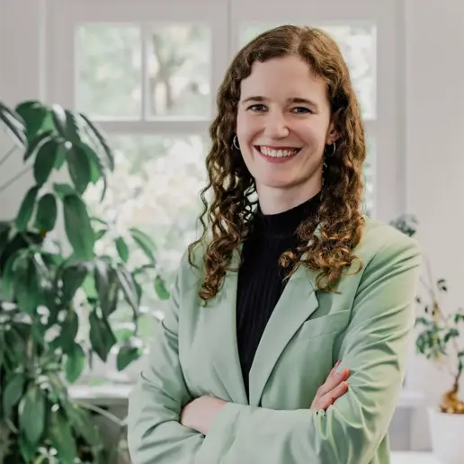 Eine Frau mit lockigem braunem Haar, die einen hellgrünen Blazer und ein schwarzes Oberteil trägt, steht lächelnd mit verschränkten Armen in einem hellen Raum mit großen Fenstern und Grünpflanzen im Hintergrund.