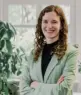 Eine Frau mit lockigem braunem Haar, die einen hellgrünen Blazer und ein schwarzes Oberteil trägt, steht lächelnd mit verschränkten Armen in einem hellen Raum mit großen Fenstern und Grünpflanzen im Hintergrund.