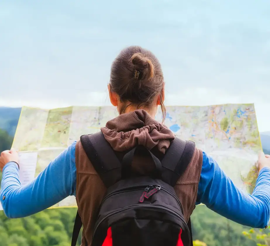 Eine Frau mit Rucksack steht im Freien, den Blick abgewandt und mit einer großen Karte in der Hand - bereit für Lernreisen - umgeben von grünen Hügeln unter einem bewölkten Himmel.
