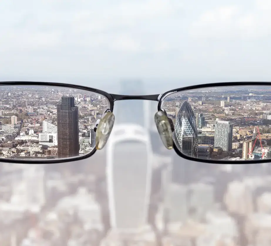 Eine Stadtlandschaft erscheint verschwommen, außer durch die Brillengläser in der Mitte, die die Gebäude scharf abbilden, was einen Perspektivenwechsel veranschaulicht und den Unterschied in der Klarheit mit und ohne Brille hervorhebt.