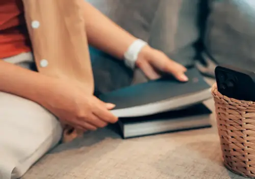 Eine Person sitzt auf einer Couch und legt ein geschlossenes Buch auf ein anderes, neben einem geflochtenen Korb mit einem Smartphone - eine Anspielung auf Digital Detox im Training. Die gemütliche Szene ist mit neutralen Kissen und sanfter Beleuchtung ausgestattet und lädt zur Entspannung abseits der Bildschirme ein.