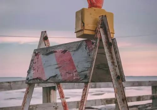 Eine verwitterte Straßensperre mit rot-weißen Streifen und einer orangefarbenen Warnleuchte steht bei Sonnenuntergang vor einem Holzzaun am Wasser und symbolisiert, dass wir Lernbarrieren abbauen müssen, um voranzukommen.