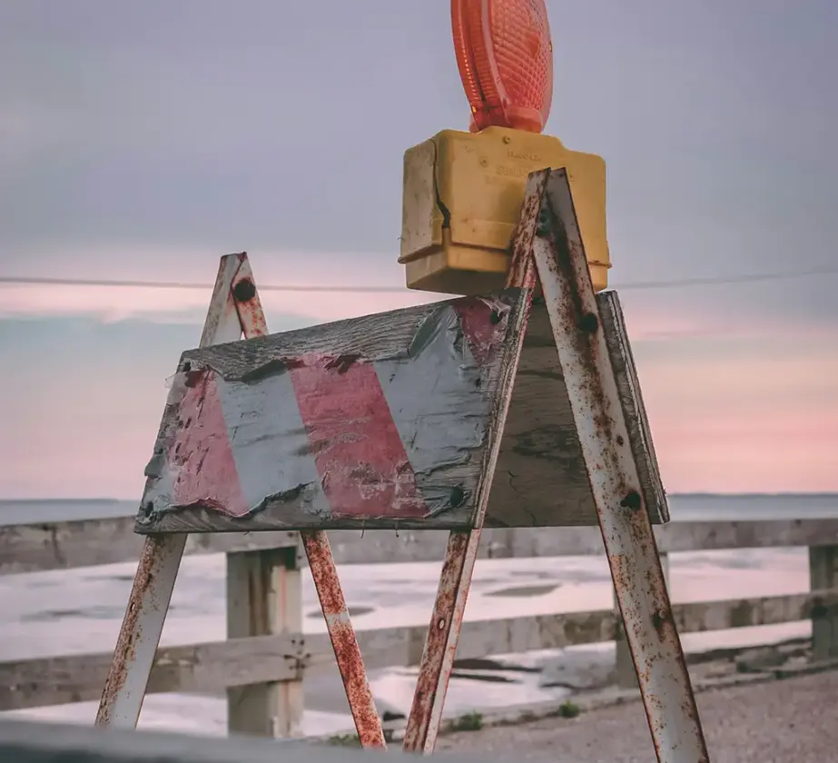 Eine verwitterte Straßensperre mit rot-weißen Streifen und einer orangefarbenen Warnleuchte steht bei Sonnenuntergang vor einem Holzzaun am Wasser und symbolisiert, dass wir Lernbarrieren abbauen müssen, um voranzukommen.