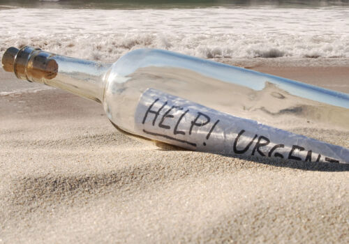 Eine Glasflasche mit einem aufgerollten Zettel mit der Aufschrift "HELP! URGENT" liegt auf dem Sandstrand in der Nähe des Ufers und symbolisiert die Notwendigkeit, Lernbarrieren in der Personalentwicklung abzubauen, während im Hintergrund die Wellen sanft rollen.