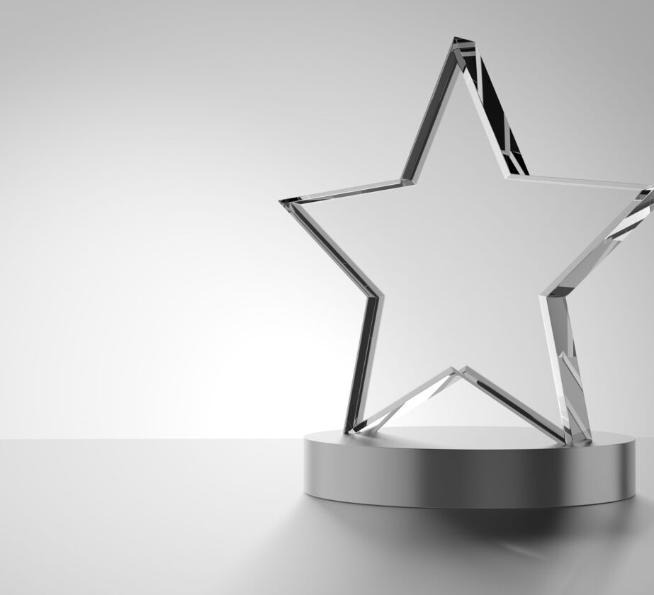 Eine sternförmige Trophäe aus Klarglas steht auf einem runden silbernen Sockel vor einem schlichten, hellgrauen Hintergrund.