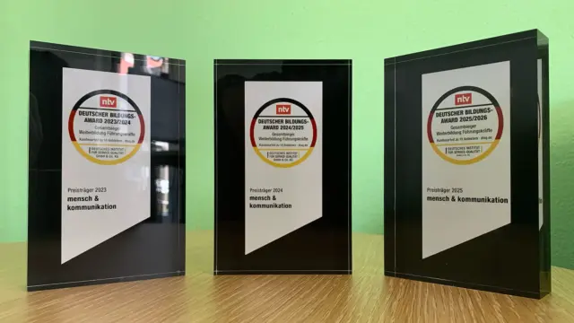 Drei gläserne Auszeichnungen mit schwarzen Sockeln und runden Etiketten mit dem Logo des Deutschen Bildungs-Awards 2023/2024 stehen auf einer Holzfläche, die Werte und Haltung bei mensch & kommunikation reflektiert, vor einer hellgrünen Wand.