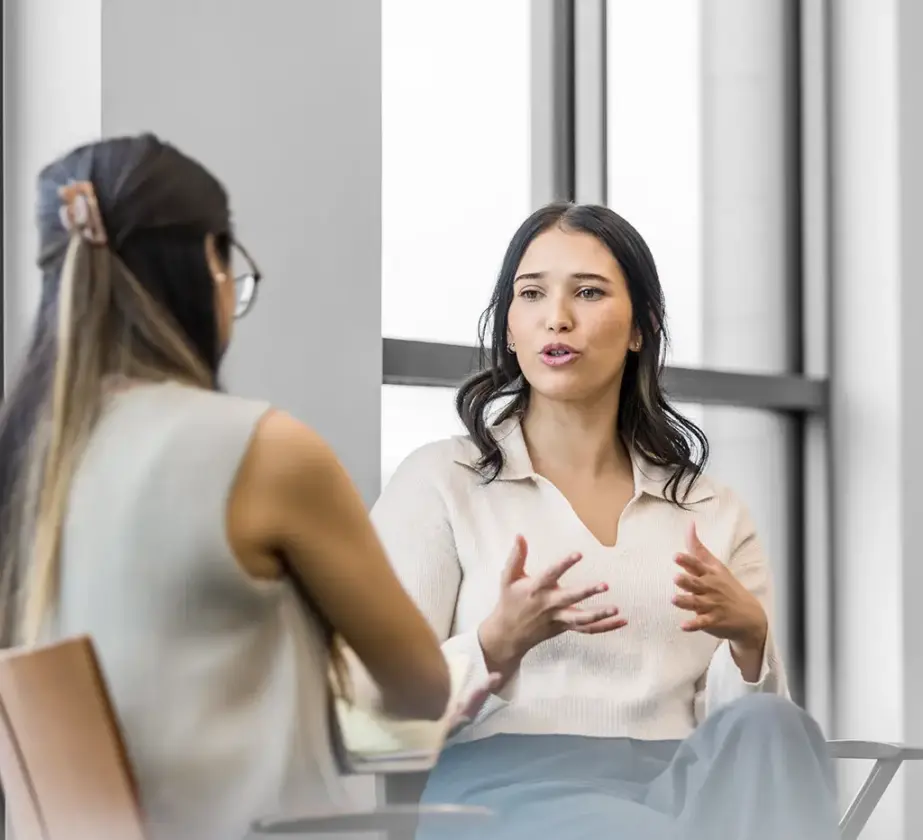Zwei Frauen sitzen in einem Haus und führen ein ernstes Coaching-Gespräch. Eine Frau, die der Kamera zugewandt ist, gestikuliert mit ihren Händen, während sie spricht, während die andere mit dem Rücken zur Kamera zuhört. Im Hintergrund sind große Fenster zu sehen.